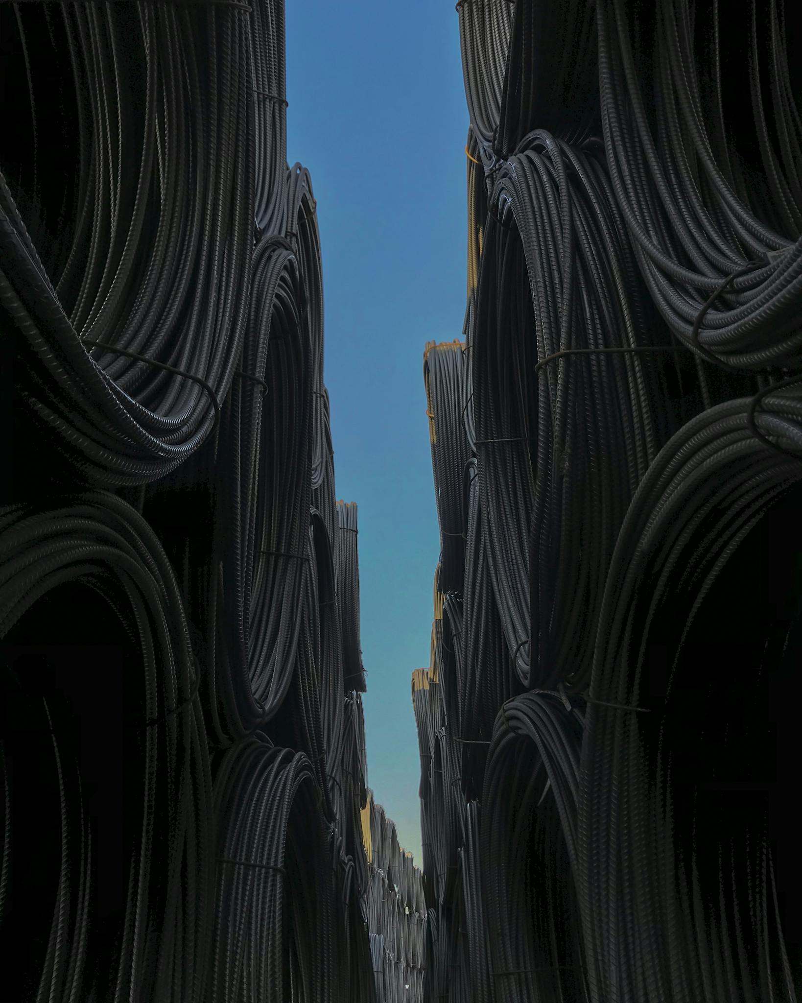 Vertical shot of piled steel bars in warehouse against a clear blue sky.